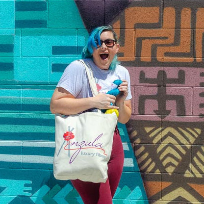 Becca is carrying an anzula tote bag in front of a graphic mural. The tote is full of yarn. She's extremely excited to have found a skein of bright blue yarn that perfectly matches her hair. 